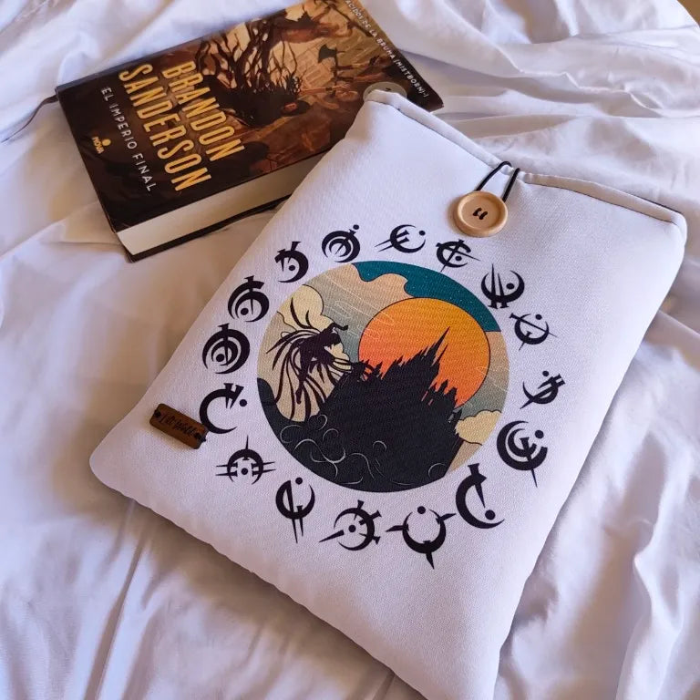 Funda de tela artesanal colocada junto a una novela de fantasía épica sobre sábanas blancas.