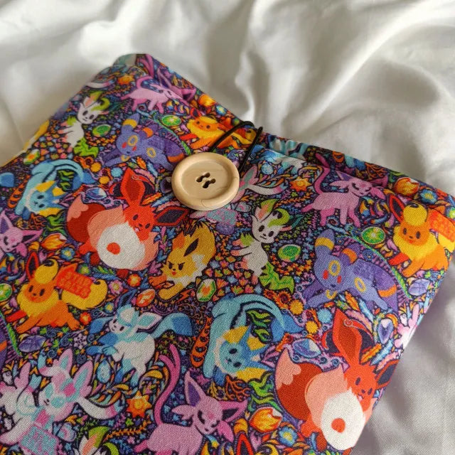 Funda de tela acolchada para libros y e-readers con diseño colorido de Eevee y sus evoluciones de Pokémon. Protección ligera y cierre con botón.