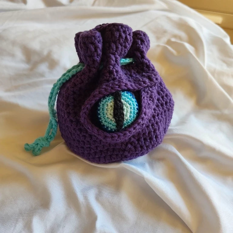 Bolsa de dados D&D de crochet en color morado y púrpura con un ojo de dragón en relieve en tonos azules.