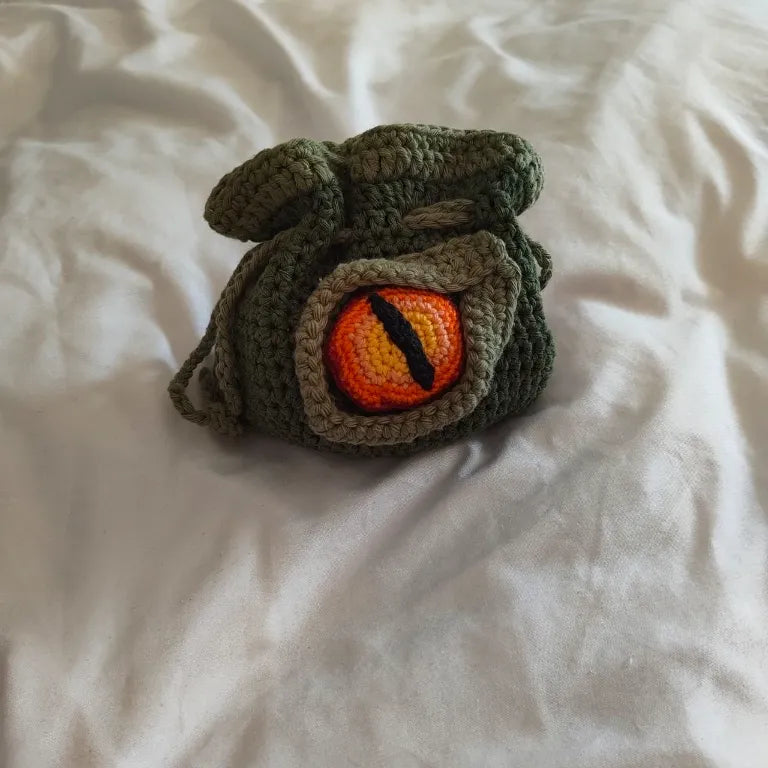 Bolsa de dados D&D de ganchillo en color verde oliva con un ojo de dragón en relieve en tonos naranja y amarillo.