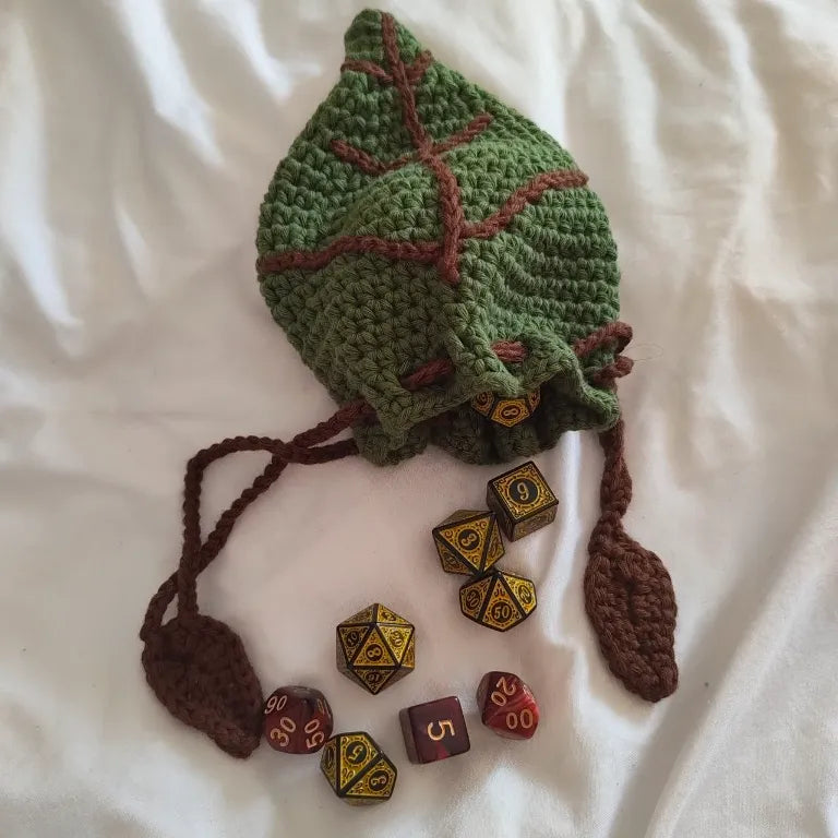 Bolsa de dados D&D verde con forma de hoja abierta en la mesa derrramando un set de dados poliédricos dorados y rojos.
