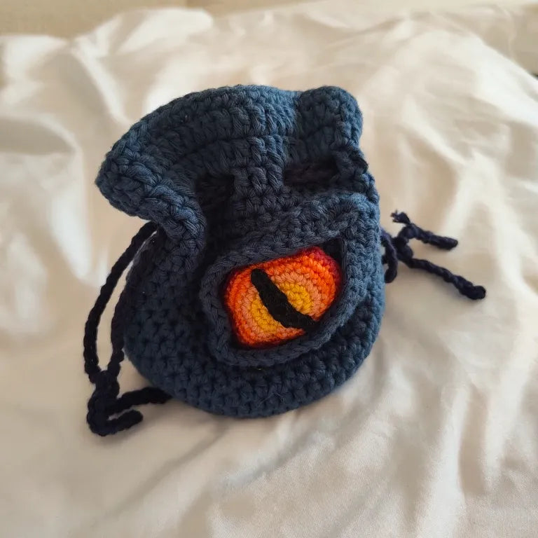 Saco para dados de rol artesanal en color azul marino con un llamativo ojo reptiliano tejido a mano.