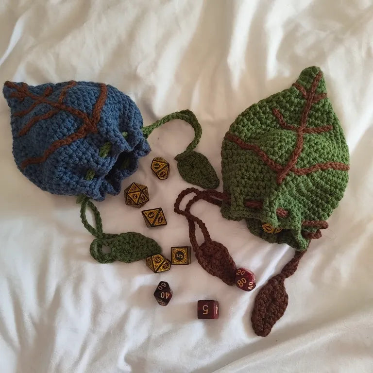 Dos bolsas para dados de rol de crochet con forma de hoja de druida, una azul y otra verde, rodeadas de dados poliédricos.