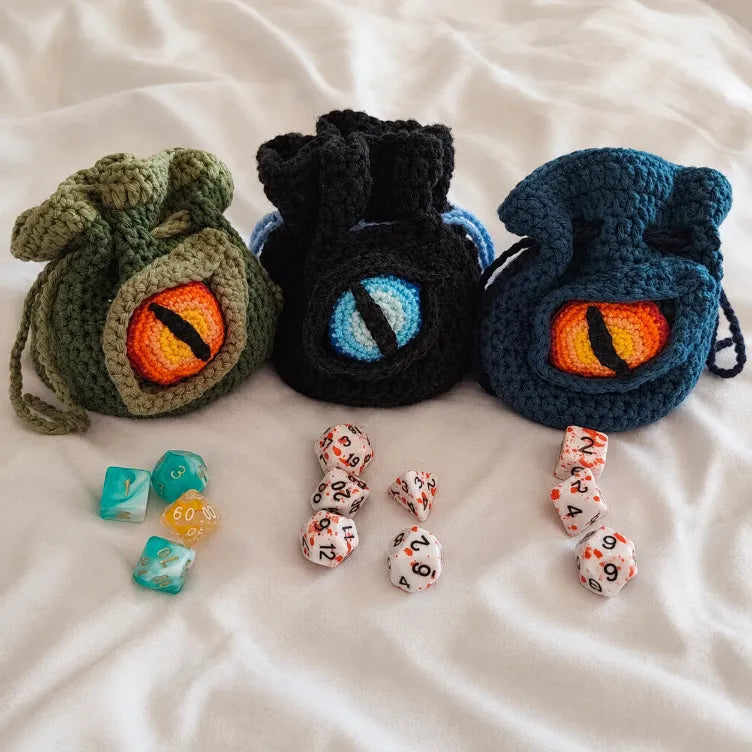 Tres bolsas para dados de rol de crochet en verde, negro y azul con ojo de dragón tejido y varios sets de dados poliédricos al frente.