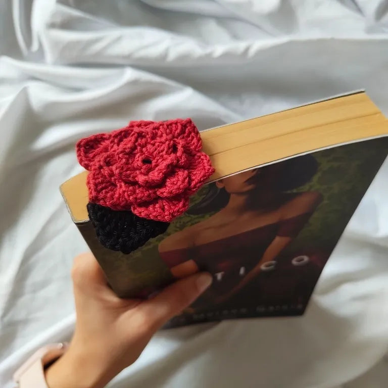 Rosa roja de crochet con hojas negras asomando sobre el borde de un libro cerrado.