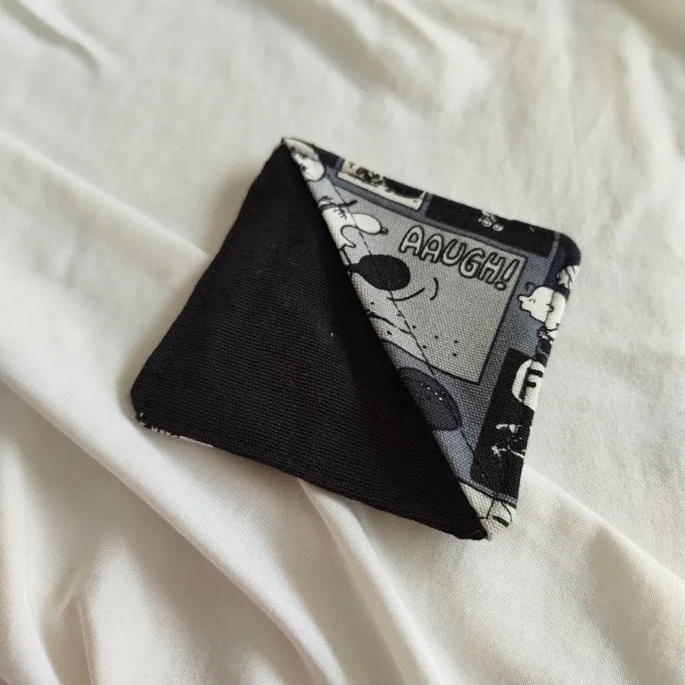 Marcapáginas esquinero triangular de tela con viñetas clásicas de la saga Peanuts sobre las páginas de un libro.